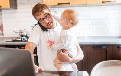 Dad working from home holding a baby who is grabbing his eyeglasses while he is standing and trying to use his laptop on the kitchen counter.