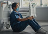 Tired health care provider wearing mask resting sitting on floor and leaning into hospital bed.